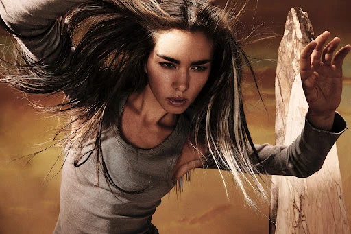 Dramatic close-up of a woman with long brown hair and grey tank top, captured with a low angle and blurred golden background.