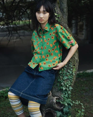 Young woman in a green floral shirt and denim skirt leaning against a tree in a park