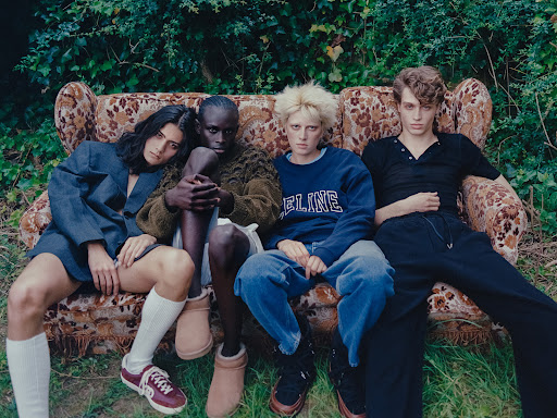 Four fashion models are sitting on a floral couch in a wooded area. They are wearing fashionable clothing and have neutral expressions.