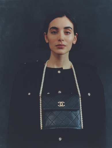 Portrait of a brunette woman wearing a black jacket and a pearl necklace with a black Chanel purse against a dark background.