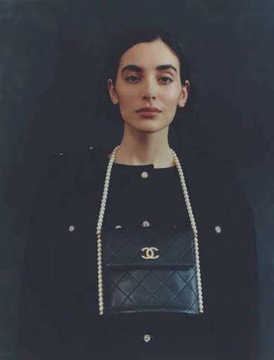Portrait of a brunette woman wearing a black jacket and a pearl necklace with a black Chanel purse against a dark background.