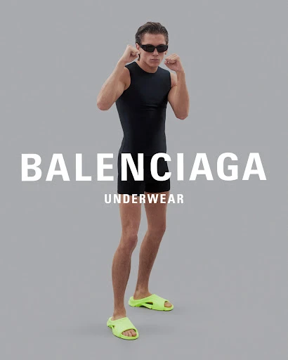 A male model poses in black athletic wear and neon slides against a gray backdrop with Balenciaga Underwear branding in front.