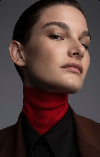 Close-up of a young adult woman with short dark hair in a black shirt, red turtleneck, and brown jacket in a studio setting.