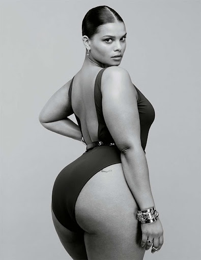Portrait of a curvy female model in a black one-piece swimsuit, wearing bracelets and earrings, looking over her shoulder in a studio setting.