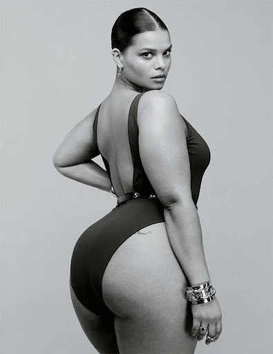 Portrait of a curvy female model in a black one-piece swimsuit, wearing bracelets and earrings, looking over her shoulder in a studio setting.