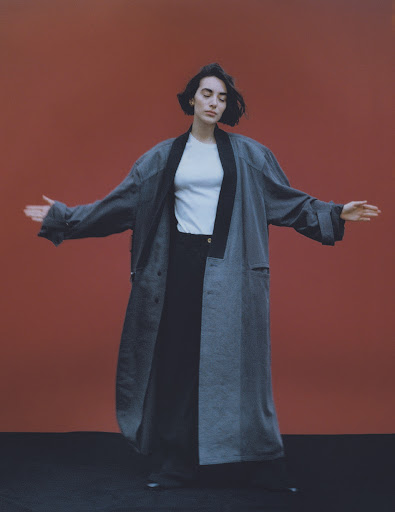 Woman with dark hair wearing a long grey coat and white t-shirt against a rust-colored backdrop with makeup by Camille Lutz.