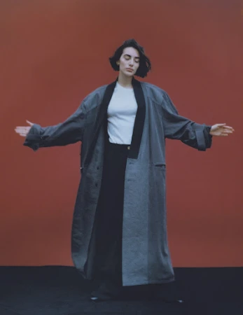 Woman with dark hair wearing a long grey coat and white t-shirt against a rust-colored backdrop with makeup by Camille Lutz.