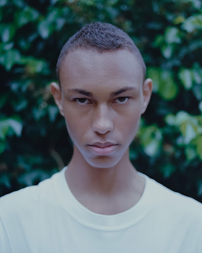 Close-up studio portrait of a young adult male with short hair and a neutral expression, wearing a white t-shirt, with a green leafy background.