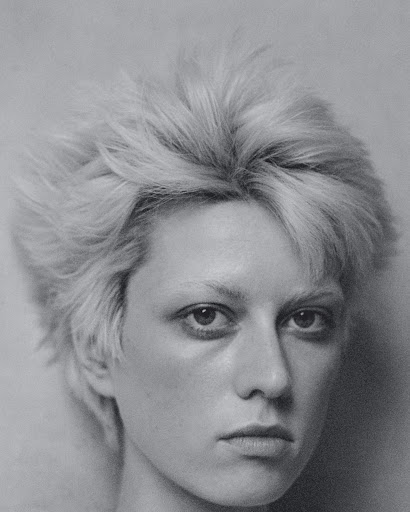 Close-up portrait of an androgynous adult, featuring a neutral expression, short blonde hair, and a monochromatic colour scheme in a photo studio.