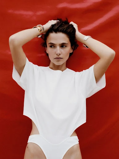 Portrait of an adult woman with dark hair and fair skin. She is wearing a white top and matching bottoms against a bright red backdrop.