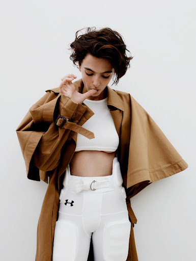 A young woman poses in a studio wearing a white crop top, white football pants, and a tan trench coat against a white backdrop.