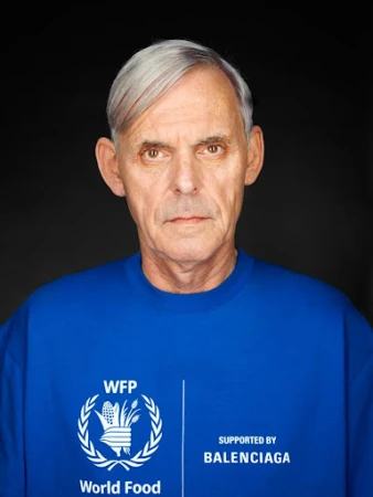 Close-up portrait of an older man with grey hair wearing a bright blue crewneck sweatshirt featuring white printed text against a dark background.