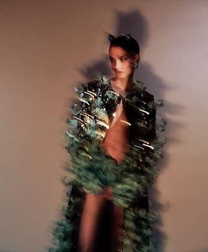 A fashionable adult female model wearing a feathered coat poses indoors against a neutral background in a glamour fashion shot.