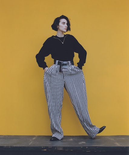 Woman with dark short hair wearing black top and striped wide trousers standing against yellow background