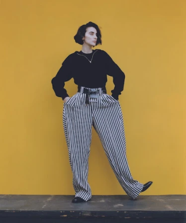 Woman with dark short hair wearing black top and striped wide trousers standing against yellow background