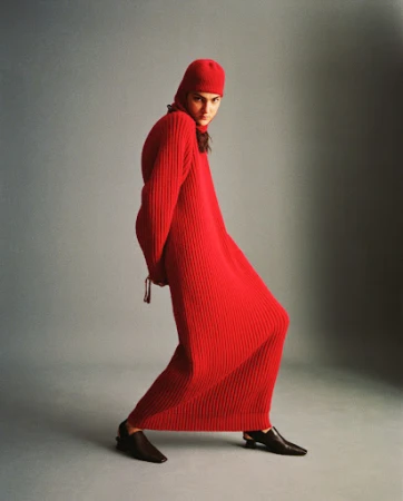 Model in a long red ribbed knit dress with matching hood and black slingback shoes against a grey studio backdrop.