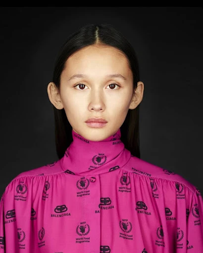 Close-up studio portrait of a young woman in a pink Balenciaga top with dark hair against a solid black backdrop, fashion-forward aesthetic.