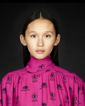 Close-up studio portrait of a young woman in a pink Balenciaga top with dark hair against a solid black backdrop, fashion-forward aesthetic.