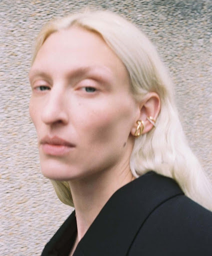 Close-up shot of a woman with light blonde hair and gold jewelry posing in front of a neutral wall wearing a black blazer.