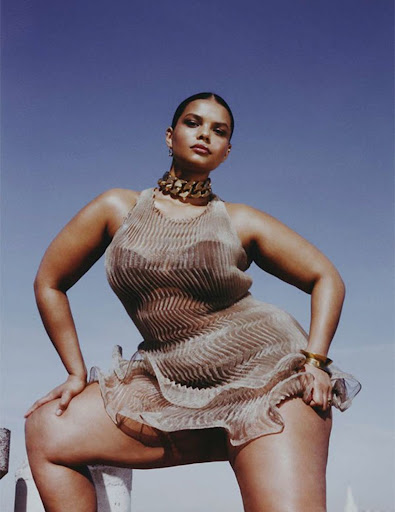 A woman poses outdoors in a shimmering, ruffled dress with a bold gold necklace under a clear blue sky, creating a fashion editorial image.