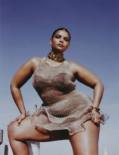 A woman poses outdoors in a shimmering, ruffled dress with a bold gold necklace under a clear blue sky, creating a fashion editorial image.