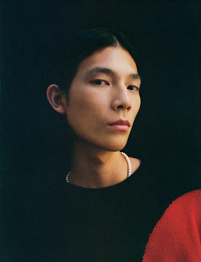Portrait of an adult Asian man wearing a black top and pearl necklace against a dark background in a studio setting.
