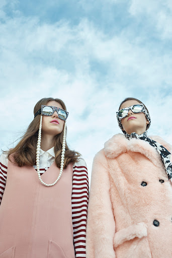 Two young women model eyewear and outerwear against a light blue sky, photographed from a low angle, emphasizing fashion and sky.