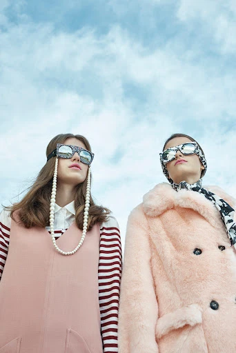 Two young women model eyewear and outerwear against a light blue sky, photographed from a low angle, emphasizing fashion and sky.
