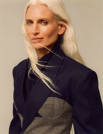 Portrait of a woman with long white hair wearing a navy blazer and plaid corset, standing in a studio with neutral background.