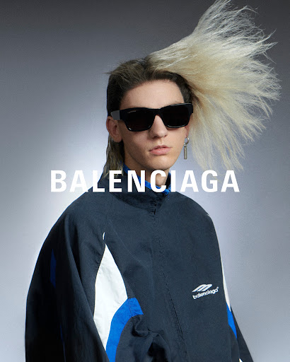 Fashion shot of a person with blond mullet wearing black sunglasses and a blue, white and black Balenciaga jacket indoors.