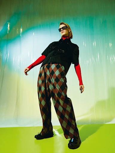 Full-length shot of a young adult with blond hair wearing sunglasses, a black shirt, patterned pants, and black shoes in a studio.