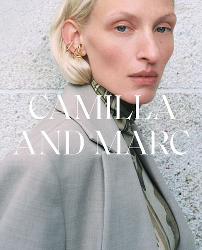 Close-up of a model in a beige jacket, showcasing gold ear jewelry and neutral makeup against a textured gray backdrop.