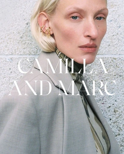 Close-up of a model in a beige jacket, showcasing gold ear jewelry and neutral makeup against a textured gray backdrop.