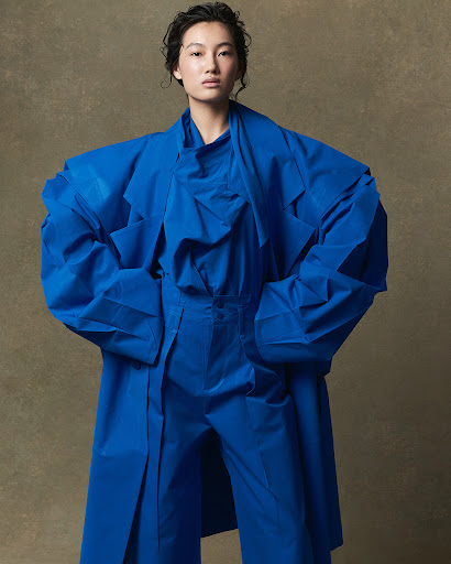 A woman poses in a blue ensemble in a studio setting, featuring oversized and stylized fashion, and a neutral backdrop.