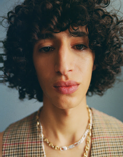 Portrait of a young adult male with curly hair wearing a patterned vest and layered chain necklaces, set against a neutral backdrop.