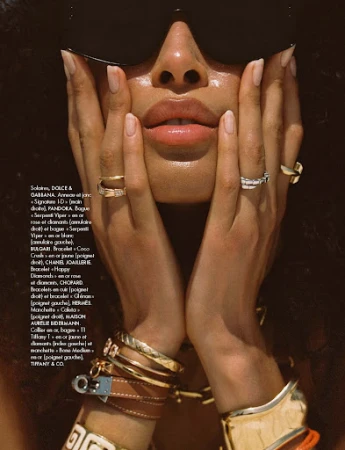 Close-up of woman framing her face with hands, wearing oversized sunglasses, multiple gold rings, and stacked bracelets.