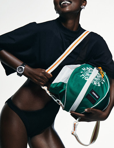 Studio shot of a Black female model wearing a black t-shirt and bottoms with a green and white duffel bag hanging from her shoulder.