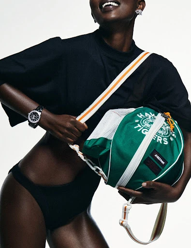 Studio shot of a Black female model wearing a black t-shirt and bottoms with a green and white duffel bag hanging from her shoulder.