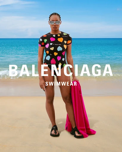 Woman in heart-print swimsuit poses on a sandy beach near turquoise ocean, holding a pink towel, for a Balenciaga swimwear ad.