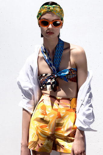 A woman with a scarf-top, head wrap, and orange sunglasses poses against a white wall, wearing printed orange shorts and an open white shirt.