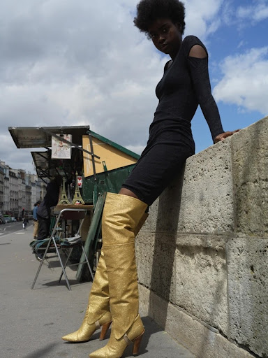 A Black woman in a short black dress and gold thigh-high boots poses on a concrete wall in an urban setting with a blue sky.
