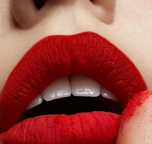 Close-up of a person's face featuring bold red lipstick on full lips, revealing a few upper teeth, and a touch of red powder.