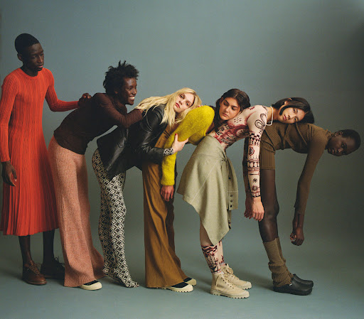Editorial fashion shot of six people of different ethnicities in fashionable outfits leaning on each other in front of a studio backdrop.