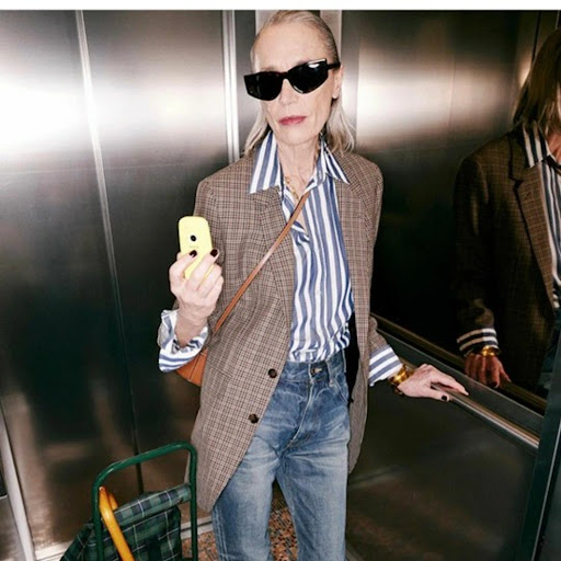 Older woman in blue jeans and a plaid blazer takes a selfie with a yellow phone in an elevator with a reflective wall.