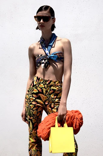 An adult female fashion model poses against a neutral wall wearing a headscarf top, patterned pants, sunglasses, and holding a handbag.