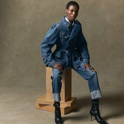 Fashionable Black woman poses in a denim jacket and jeans, seated on a wooden block, paired with stylish black leather boots in studio.