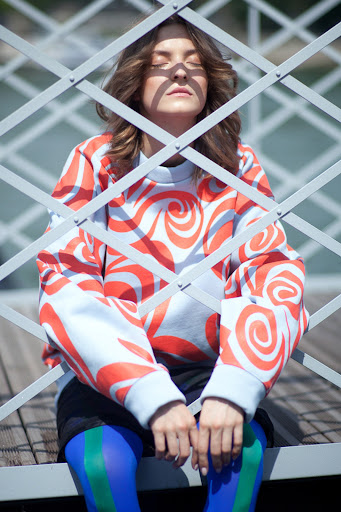 A young adult woman with wavy brown hair, wearing a patterned sweatshirt and colorful tights, poses behind a metal fence.