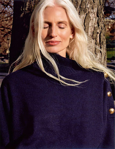 Mature woman with long blonde hair and closed eyes wearing a navy turtleneck sweater outdoors against a tree trunk.