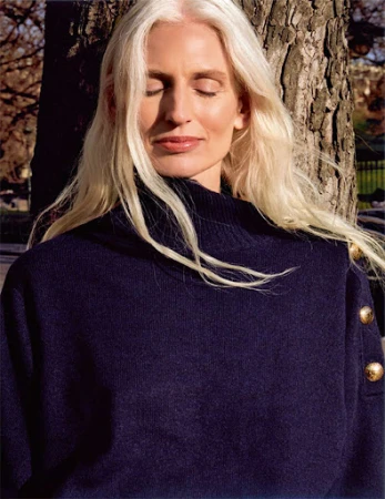 Mature woman with long blonde hair and closed eyes wearing a navy turtleneck sweater outdoors against a tree trunk.