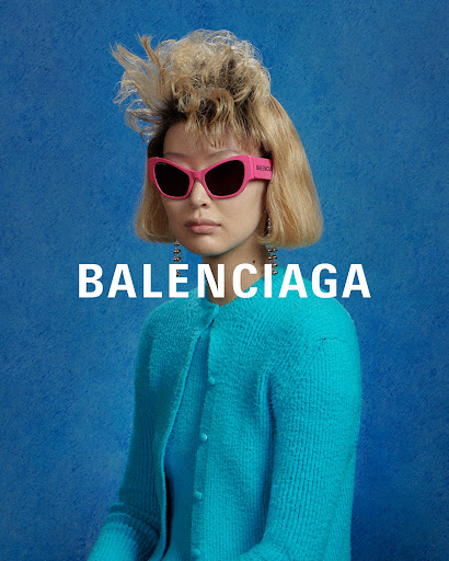 Portrait of a fair-skinned woman wearing pink sunglasses and a turquoise cardigan, set against a blue background with 'BALENCIAGA' text.